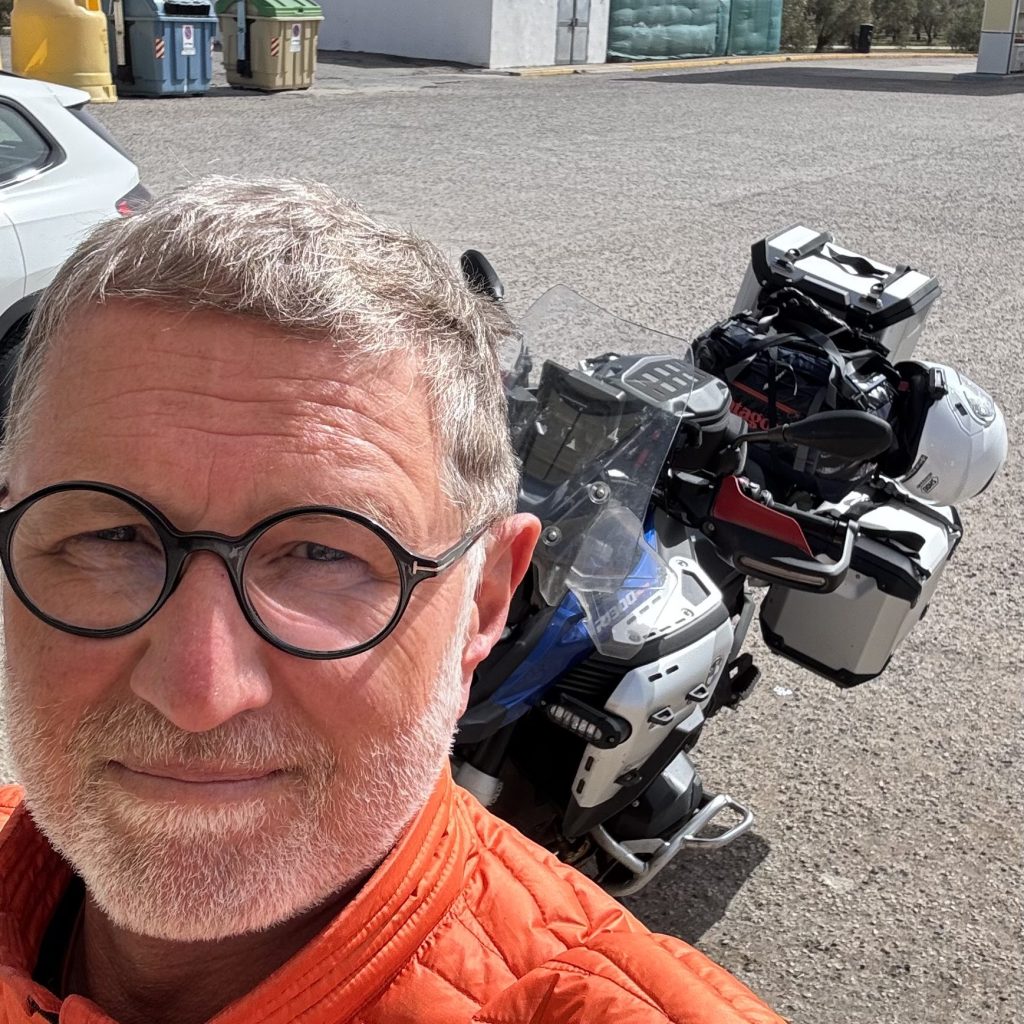 En man med grått hår och rundade glasögon tar en selfie framför en motorcykel med bagageutrustning.